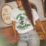 A woman in glasses and a white Austin FC vintage t-shirt from the Verde Collection, featuring a cartoon monkey, soccer ball, and the text “SOMOS VERDES” and “Howler Brothers,” stands indoors with one hand on her waist and long brown hair over her shoulder.
