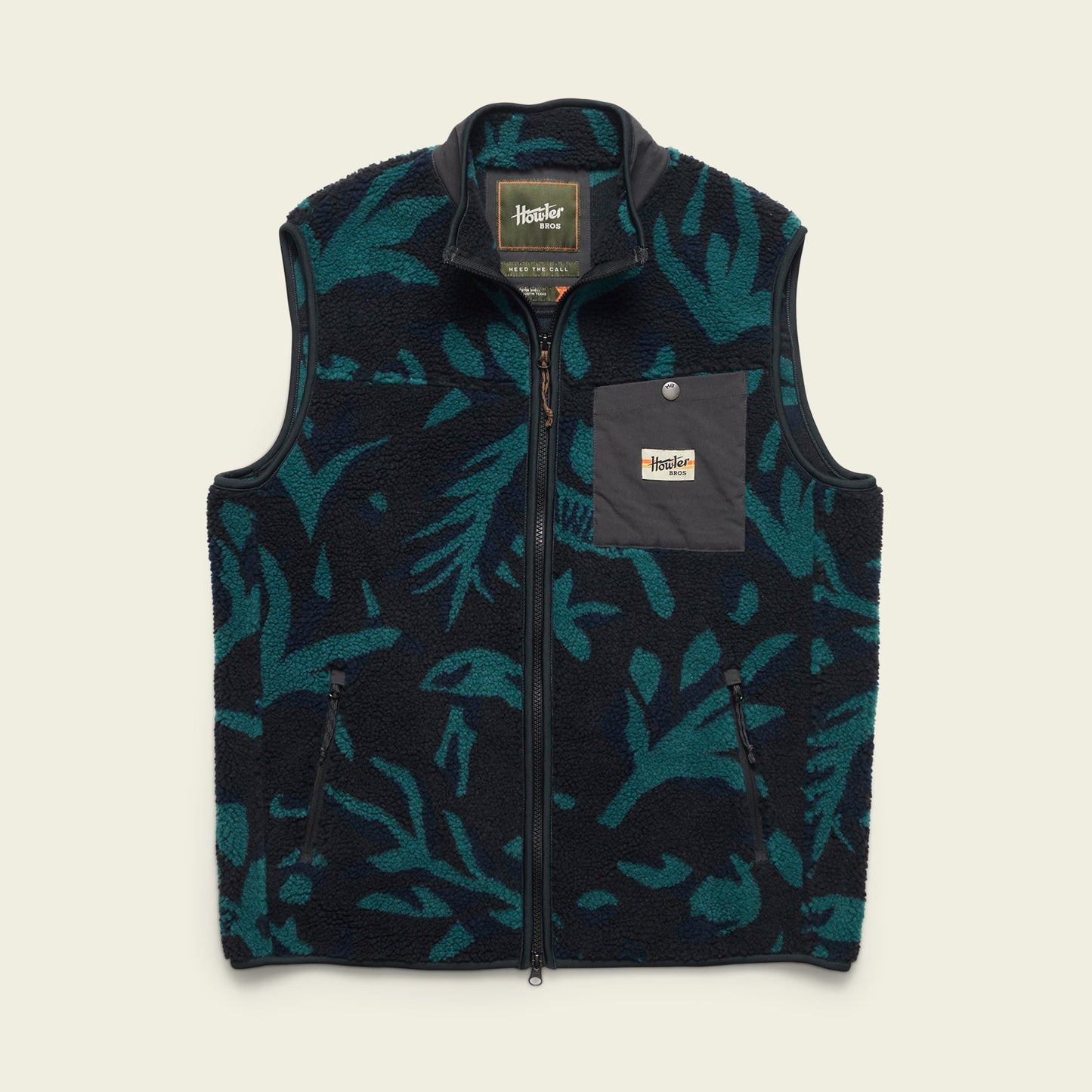The Chisos Fleece Vest is a lightweight vest featuring a dark blue and teal leafy pattern, high pile fleece, a full front zipper, high collar, and a gray chest pocket with a rectangular “Howler Bros” logo patch.