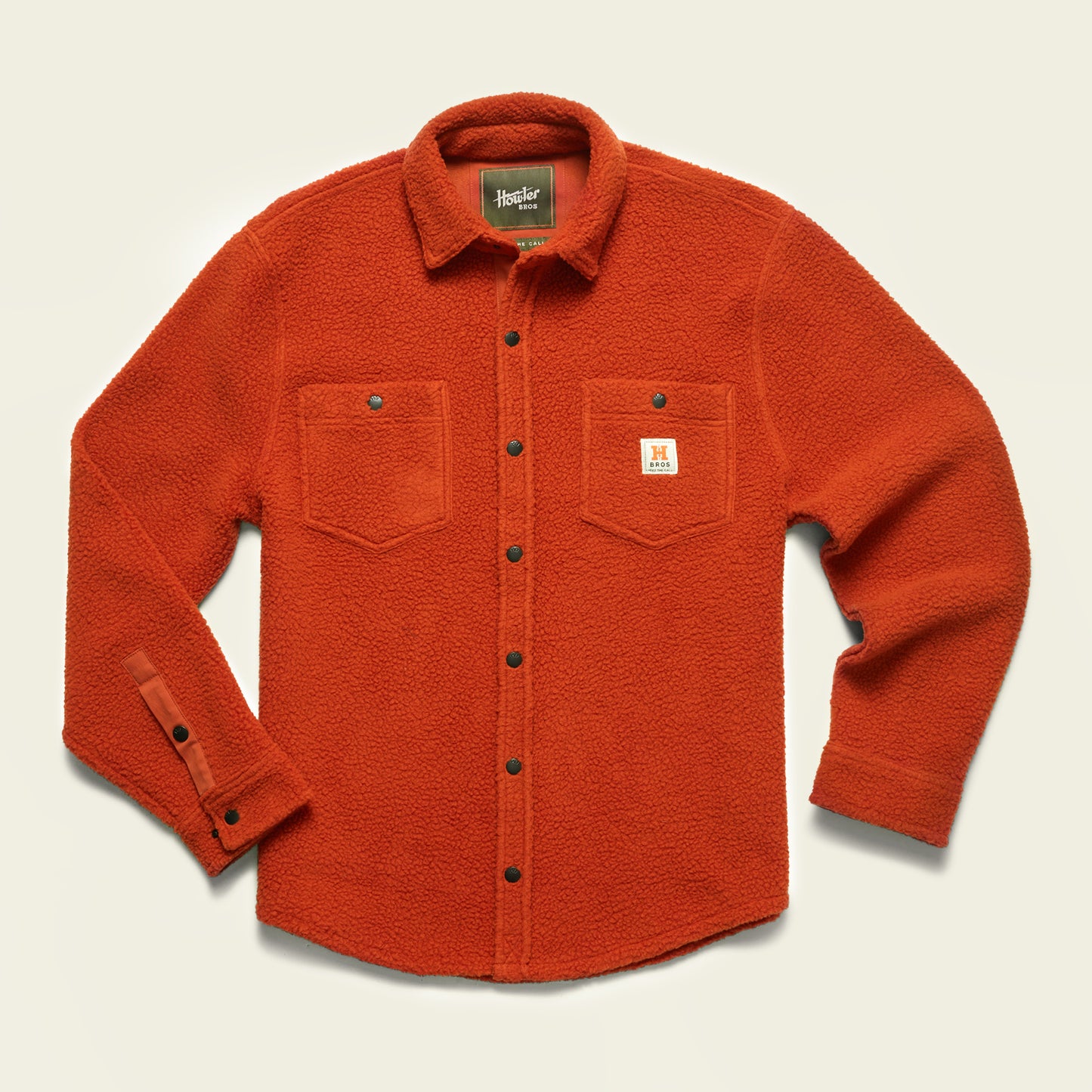 An orange fleece overshirt with two chest pockets, metal snap buttons, and a small white patch on the left pocket displaying an H logo. The shirt has a casual, textured appearance perfect for layering as a casual jacket.