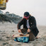 A person in a casual jacket and cap sits on a sandy beach, slicing cooked meat on a wooden board. Beach houses and cliffs appear in the background, while another person with a drink relaxes in the foreground.