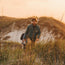 A man with curly hair, wearing a green jacket and khaki pants, hikes through tall grass on sandy dunes at sunset, carrying a backpack and a water bottle, layered in a cozy fleece pullover for cold weather.