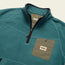 A teal fleece pullover designed for cold weather, featuring a black quarter zip, black collar trim, and an olive green chest pocket with a snap button and Howler Bros logo patch.