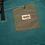 Close-up of a teal Fleece Pullover with an olive green front pocket. The pocket has a snap button and a stitched patch label reading Howler Bros, with an orange and yellow stripe—perfect for cold weather comfort.