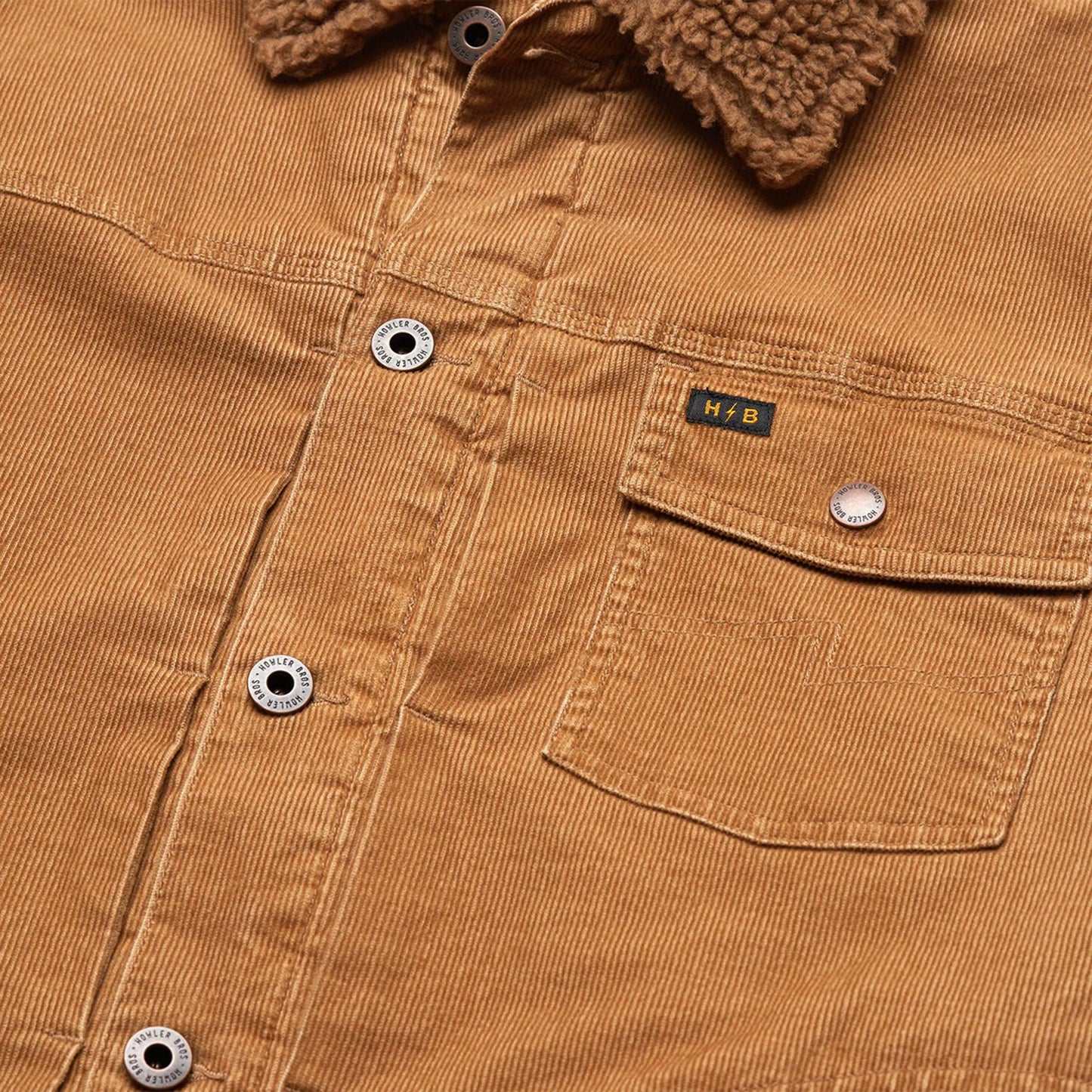 Close-up of a tan corduroy jacket, known as the Fuzzy Depot Jacket, featuring metal buttons, a brown sherpa collar, chest pocket, sherpa lining for warmth, and a small yellow “H.B” label above the pocket.