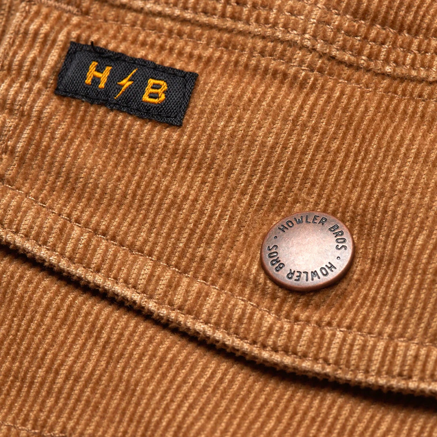Close-up of brown corduroy jacket fabric with a black and yellow “H//B” label and a metal button engraved with “HOWLER BROS.”—a signature detail on the Fuzzy Depot Jacket.