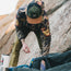 A person in a camouflage jacket and breathable tech hat sorts through outdoor gear beside a rock wall. Their face is obscured by the cap, and quick drying camping equipment is visible in the foreground.