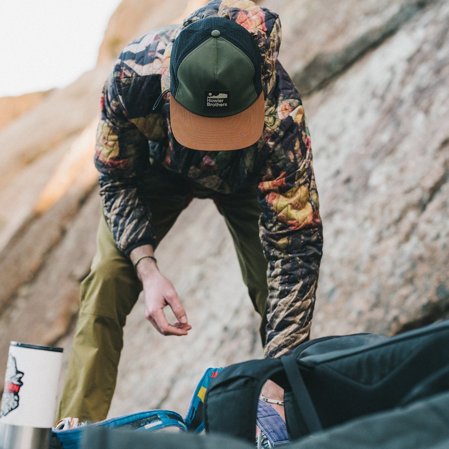 A person in a camouflage jacket and breathable tech hat sorts through outdoor gear beside a rock wall. Their face is obscured by the cap, and quick drying camping equipment is visible in the foreground.