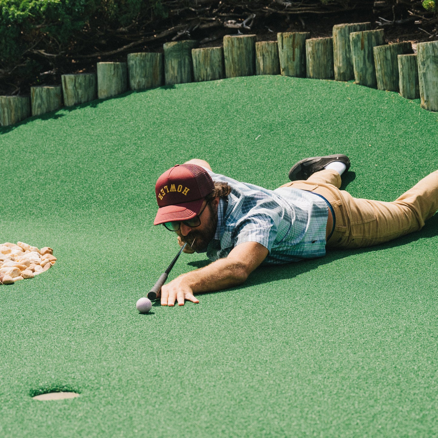 A man lies on his stomach on artificial turf at a mini-golf course, stretching out to putt a golf ball with his club held in his mouth. He wears a vintage-inspired hat, sunglasses, a plaid shirt, and khaki pants.