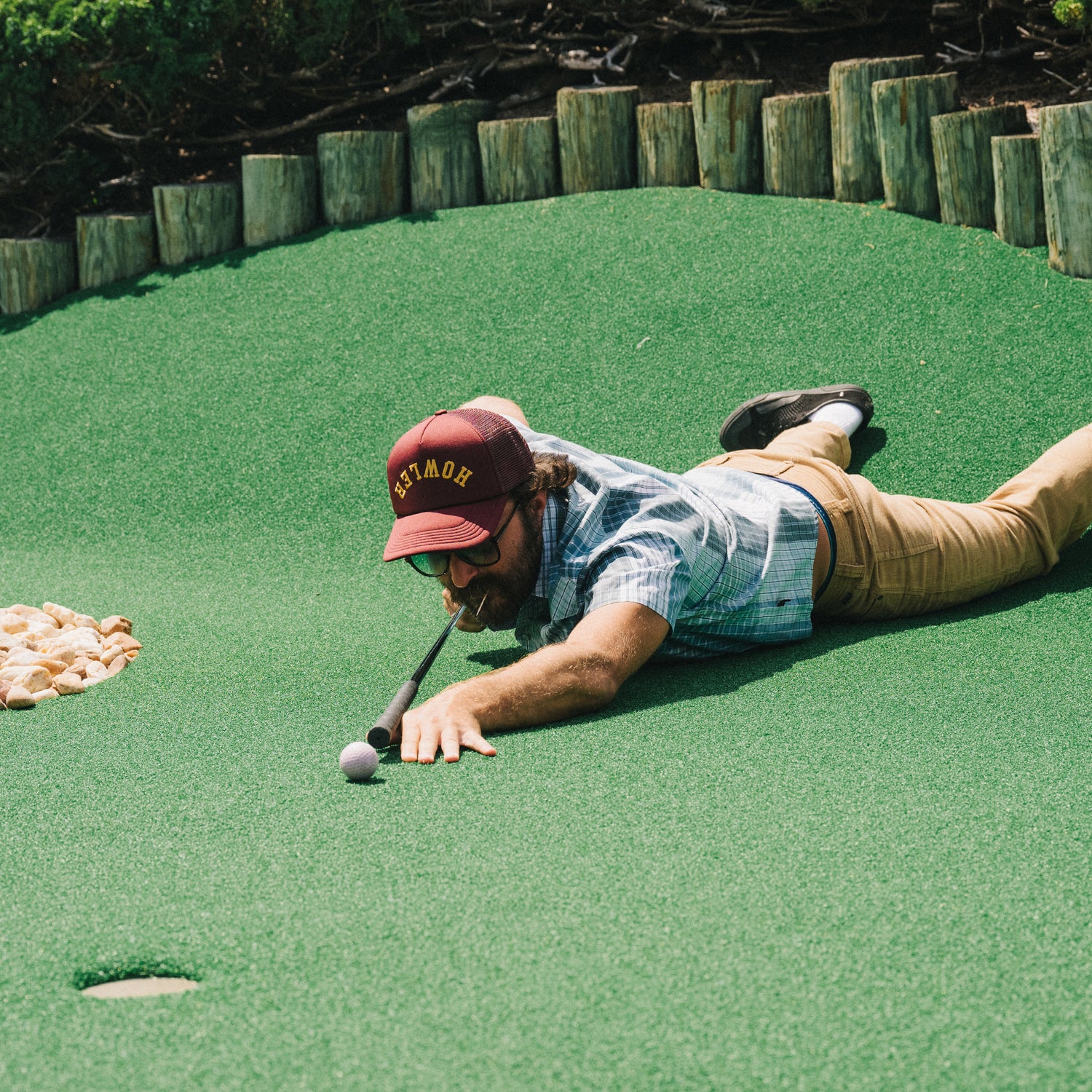 A man lies on his stomach on artificial turf at a mini-golf course, stretching out to putt a golf ball with his club held in his mouth. He wears a vintage-inspired hat, sunglasses, a plaid shirt, and khaki pants.