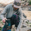 A person in a gray sweater and cap with an adjustable snapback closure prepares a camping stove on rocky ground near a river, holding a blue container and looking down at their task.