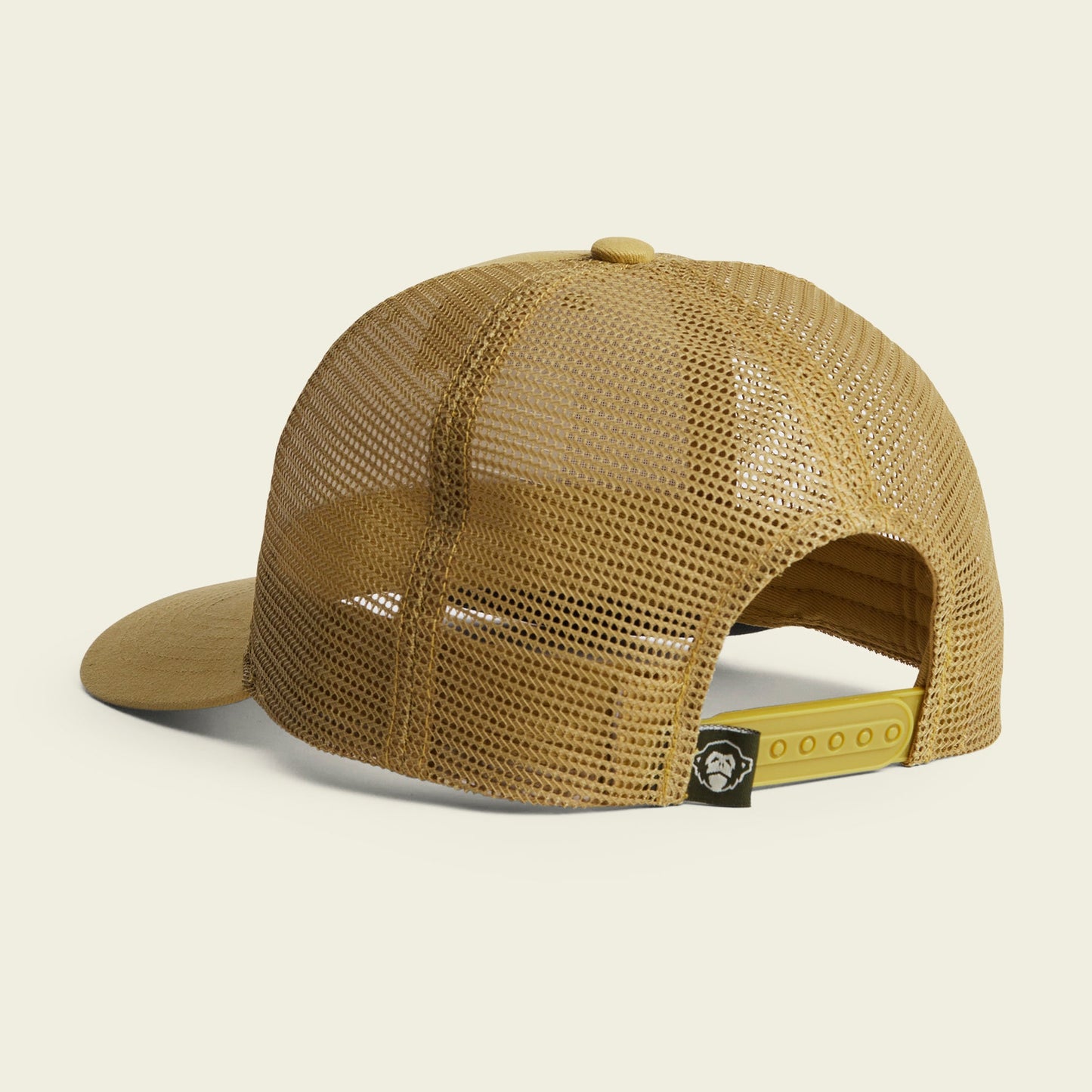 Tan mesh trucker hat seen from the back, featuring an adjustable snapback closure for a custom fit and a small black tag with a white monkey face logo—signature Howler artwork detail.