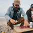 A man in a denim jacket kneels on a sandy beach, seasoning raw meat on a wooden board. Another casually dressed person sits nearby, with the ocean and sky stretching in the background.