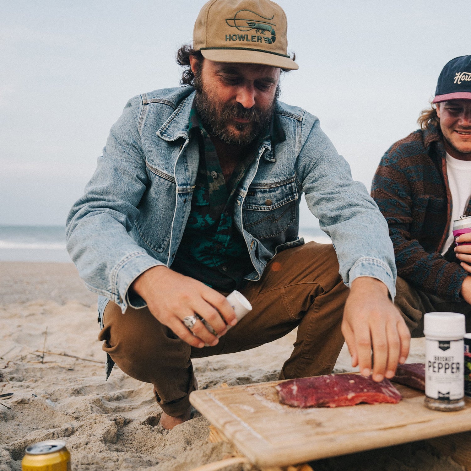 A man in a denim jacket kneels on a sandy beach, seasoning raw meat on a wooden board. Another casually dressed person sits nearby, with the ocean and sky stretching in the background.