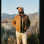 A man in a tan corduroy jacket, white pants, and a baseball cap stands outdoors among dry bushes with mountains in the background under a clear sky.