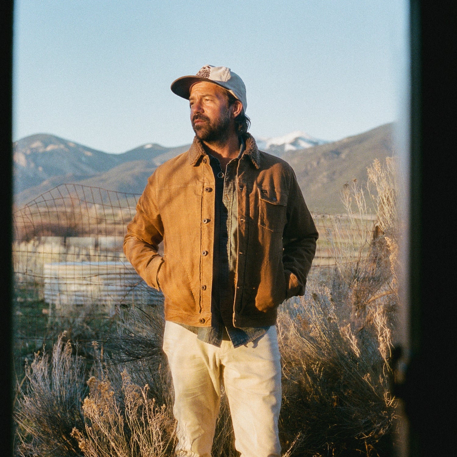 A man in a tan corduroy jacket, white pants, and a baseball cap stands outdoors among dry bushes with mountains in the background under a clear sky.