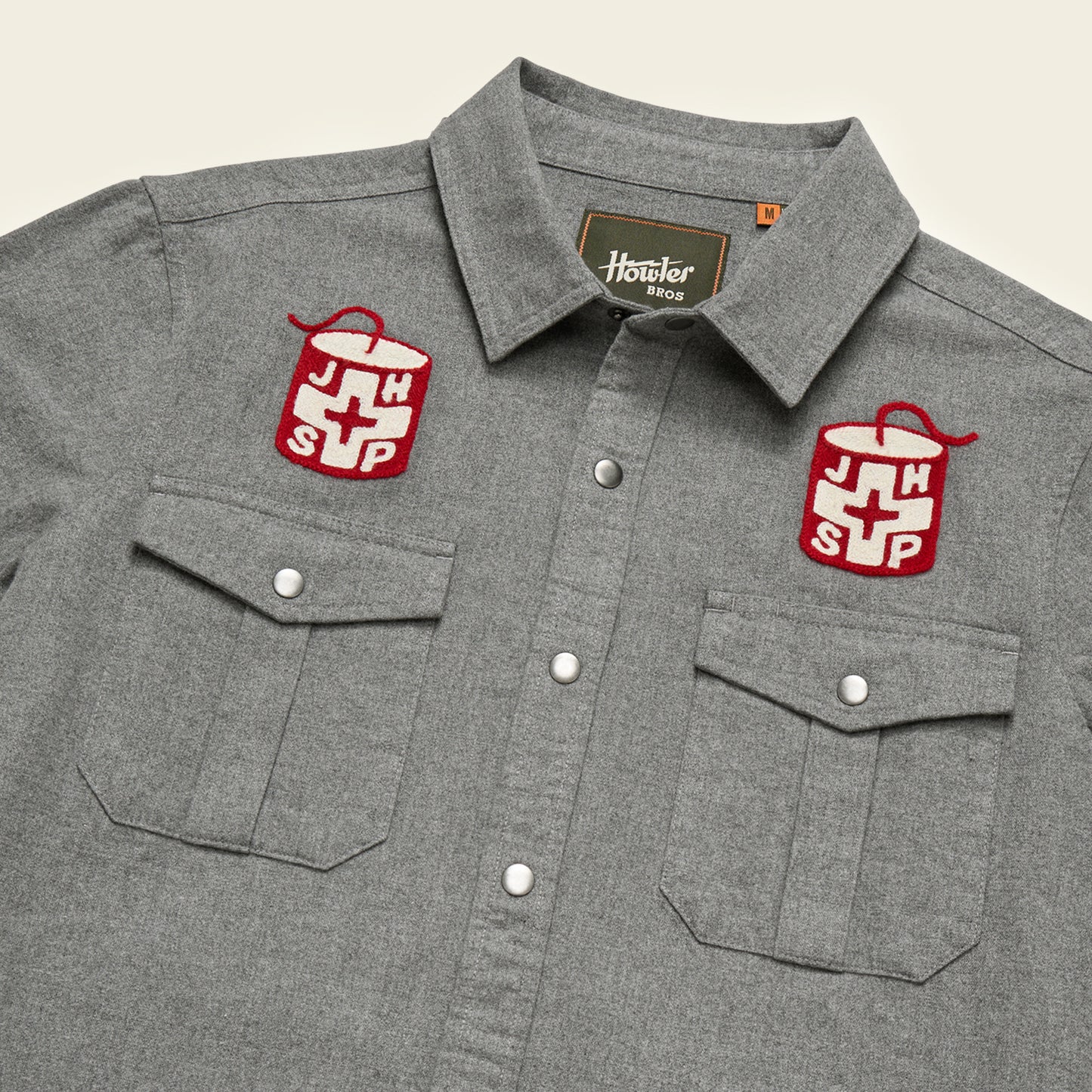 A gray Gaucho Snapshirt with two chest pockets and embroidered red and white Jackson Hole Ski Patrol patches above each pocket, featuring a cross design. The label inside the collar reads Howler Bros.