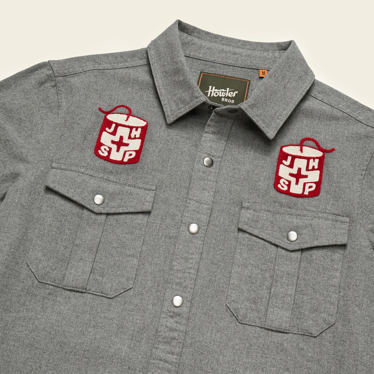 A gray Gaucho Snapshirt with two chest pockets and embroidered red and white Jackson Hole Ski Patrol patches above each pocket, featuring a cross design. The label inside the collar reads Howler Bros.