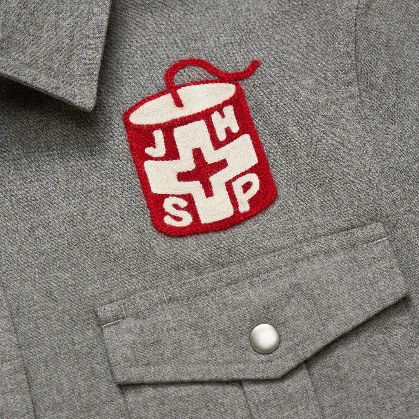 A gray flannel shirt with a buttoned pocket and a patch showing a red can with a white cross and the letters J, H, S, and P—perfect for Jackson Hole Ski Patrol fans.