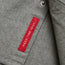 A close-up of gray flannel shirt fabric with a red stitched tag that reads HEED THE CALL in white capital letters. There is also a nearby metal snap button, hinting at the quality of a Gaucho Snapshirt.