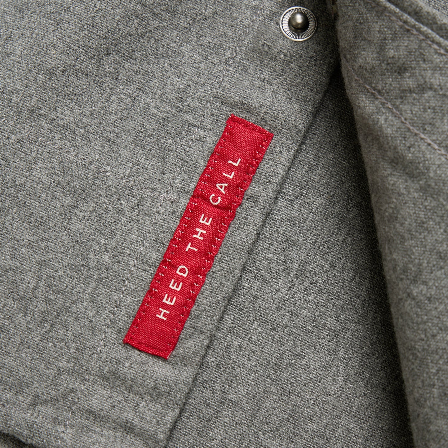 A close-up of gray flannel shirt fabric with a red stitched tag that reads HEED THE CALL in white capital letters. There is also a nearby metal snap button, hinting at the quality of a Gaucho Snapshirt.