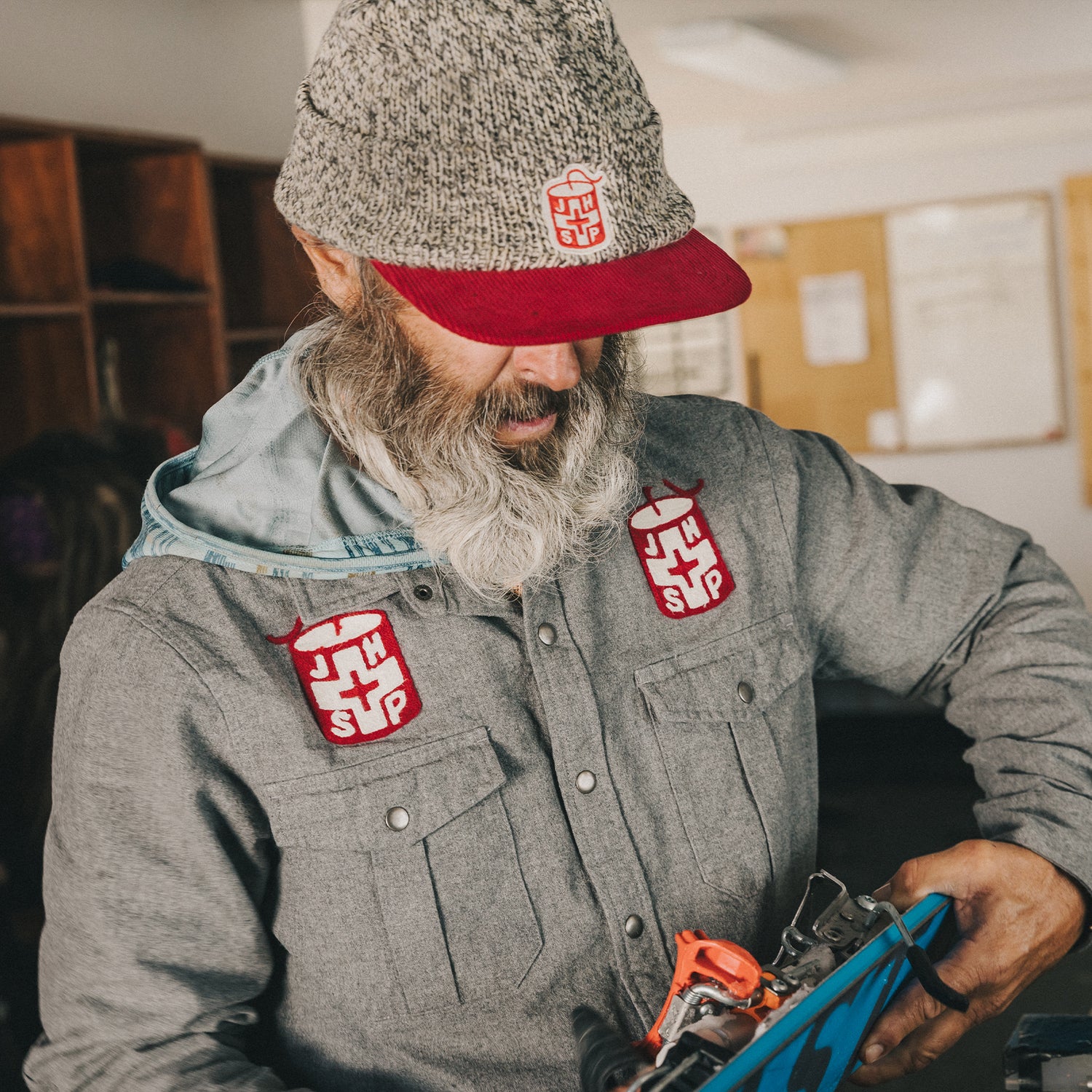 A bearded man in a gray flannel shirt and knit hat with red and white patches inspects ski equipment indoors, with a classroom-style background and bulletin boards behind him.