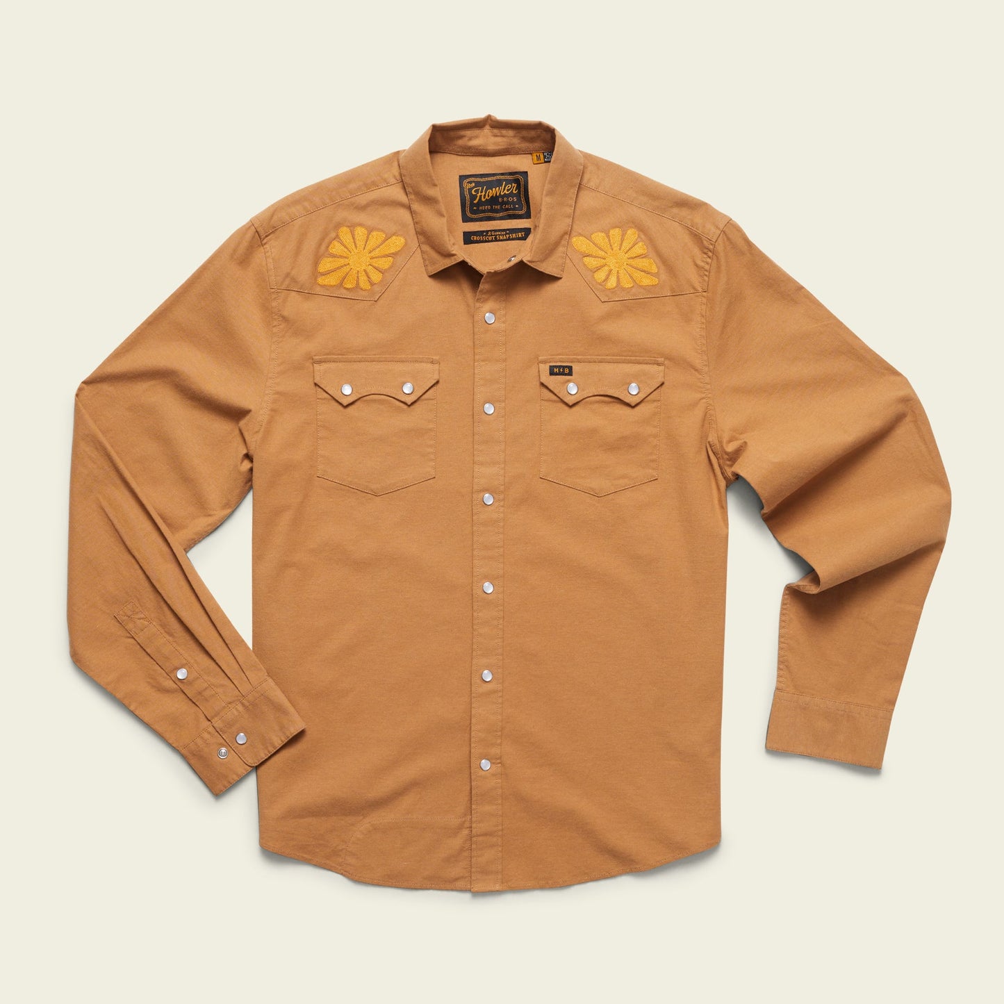 A long-sleeve, button-up tan shirt with pearl snaps, two front chest pockets, and vintage inspired motif embroidery in yellow on the shoulders, displayed on a light background.