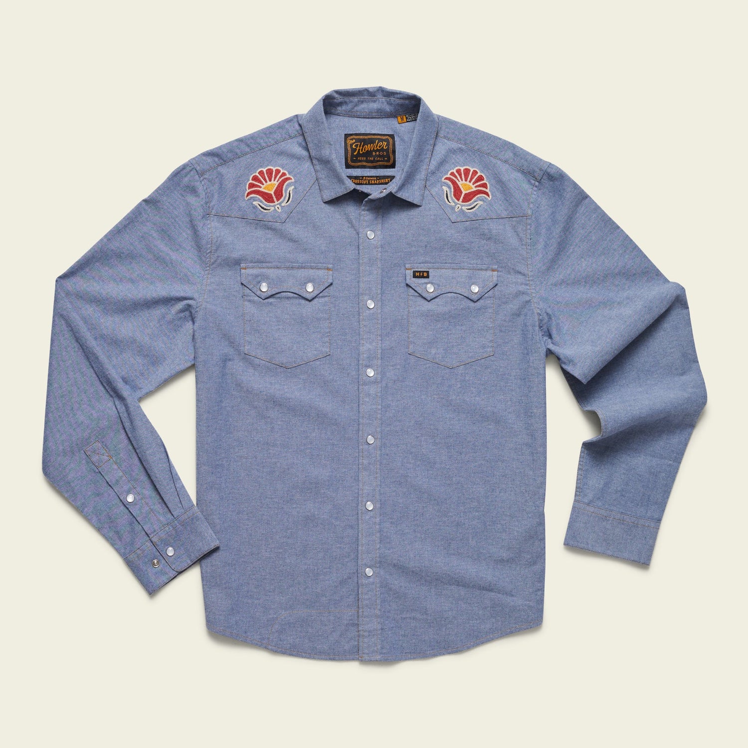 Light blue long-sleeve button-up shirt crafted from stretch chambray, featuring two chest pockets and pearl snaps. Red and white vintage inspired motif embroidery decorates each shoulder near the collar.