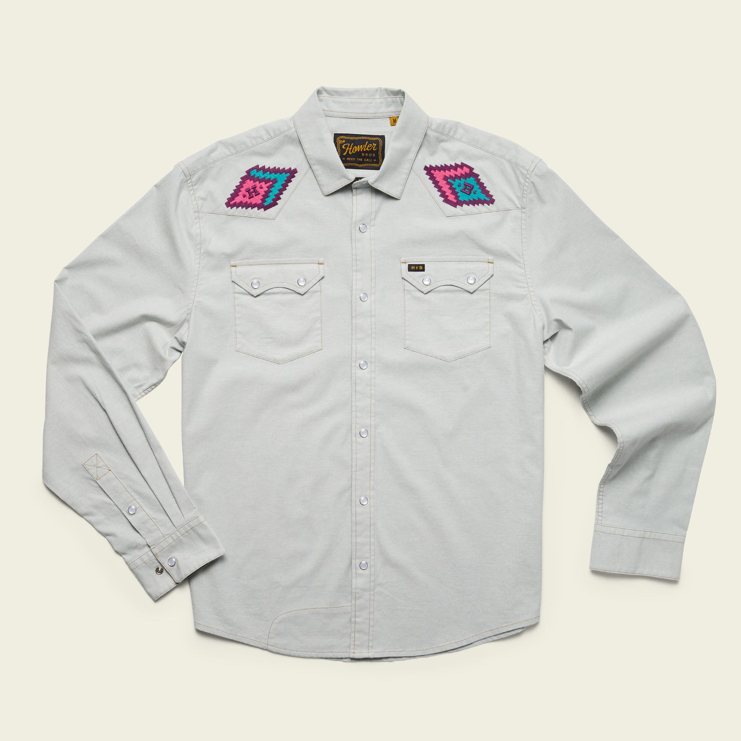 A light gray, long-sleeve stretch chambray button-up shirt with pearl snaps, two chest pockets, and colorful, vintage inspired motif embroidery in pink, purple, and blue on each shoulder. The shirt is laid flat on a cream background.