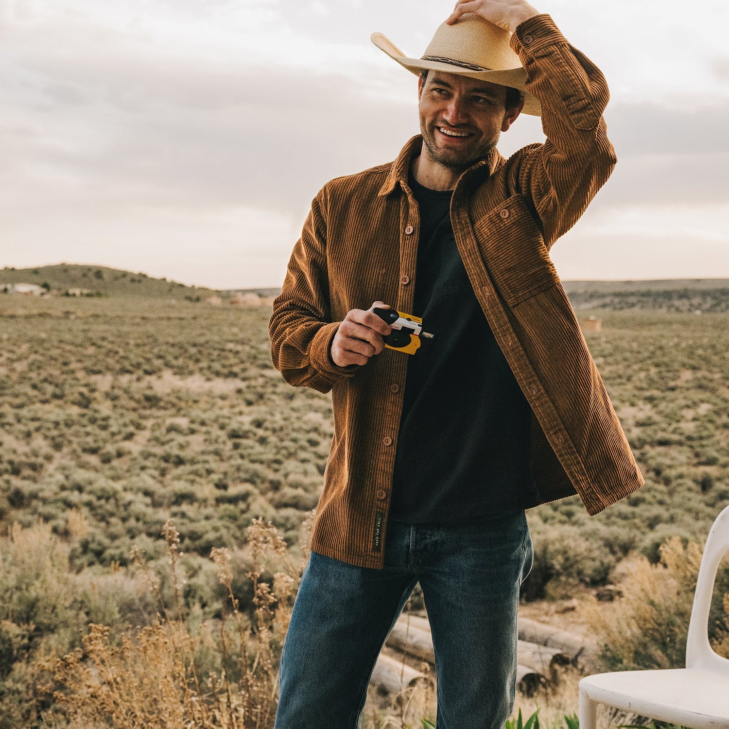 A smiling man in a brown jacket and jeans stands outdoors in a field, holding a small toy and adjusting his cowboy hat with one hand. His Iquitos Overshirt adds rugged style against the open countryside and cloudy sky.