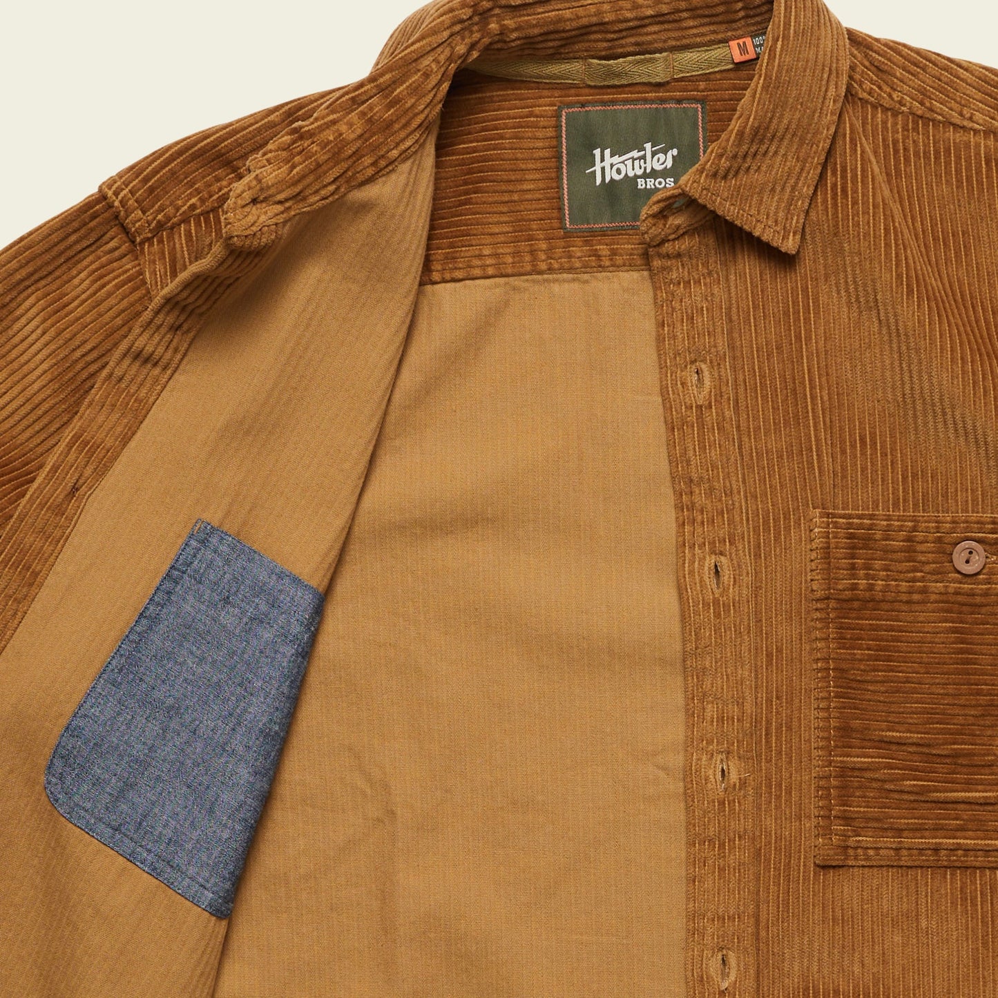 A brown wide wale corduroy travel shirt with the front open, revealing a blue inner pocket. The Iquitos Overshirt features a chest pocket and a Howler Bros label sewn inside the collar.