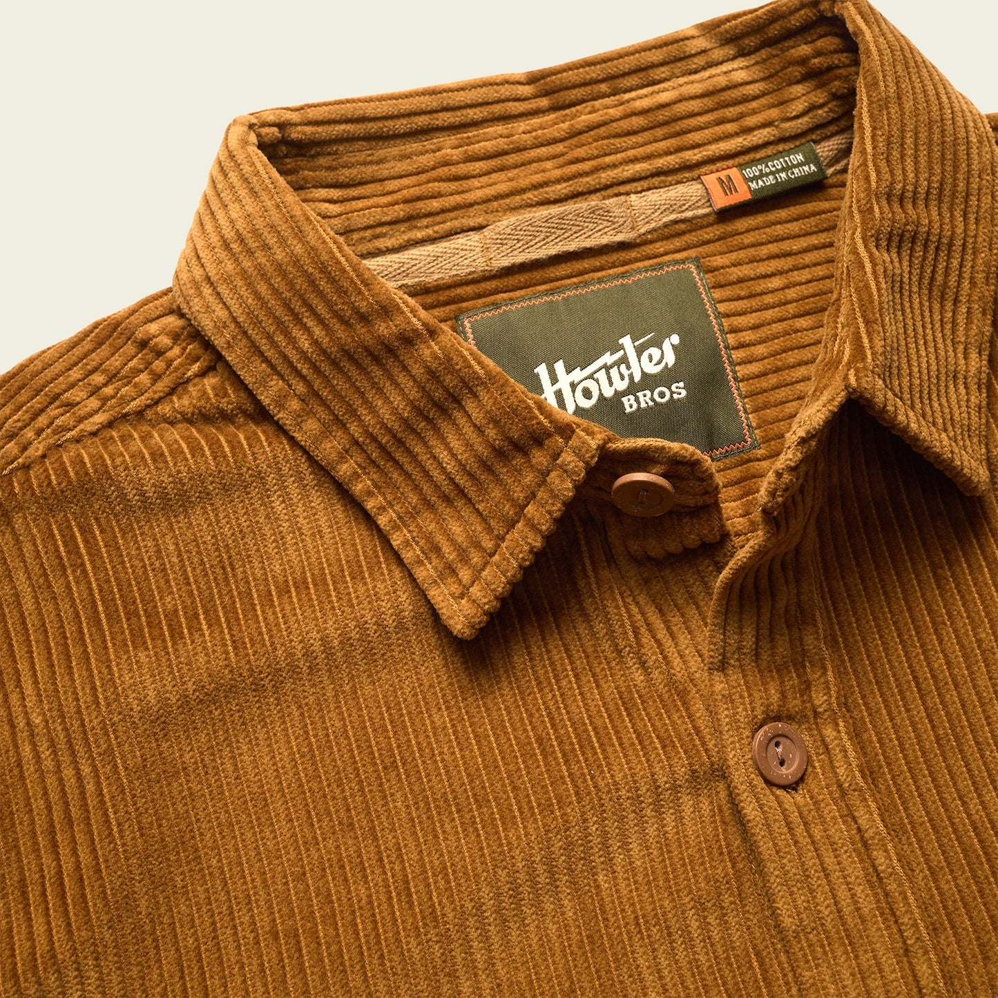 A brown wide wale corduroy button-up shirt with a collar, featuring two labels inside the neck area: one says Howler Bros, and the other indicates it is a medium size and made in Vietnam.