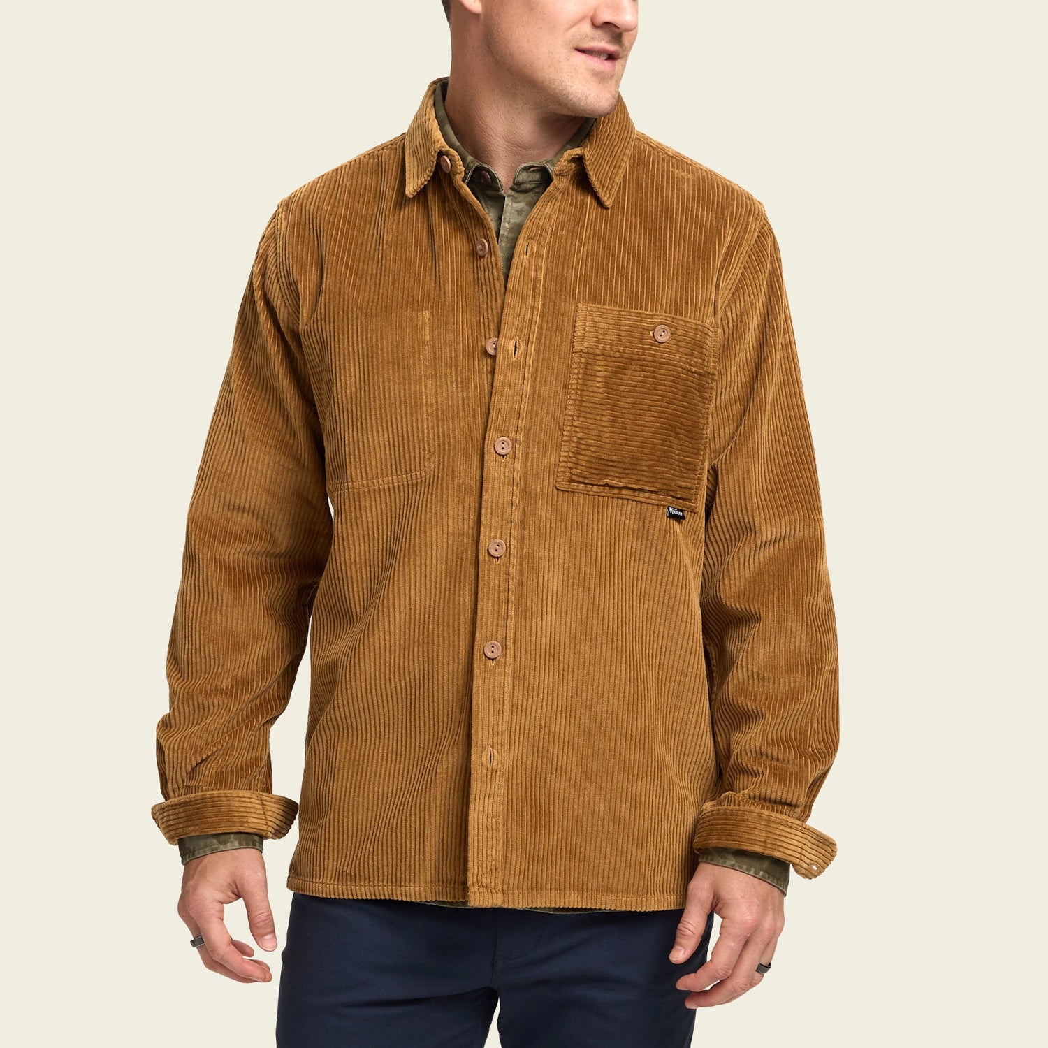 A man wearing a tan wide wale corduroy Iquitos Overshirt with a front chest pocket, layered over a green shirt and dark pants. He stands with his head turned slightly to the side—a perfect travel shirt look.