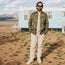 A man wearing sunglasses, a camouflage jacket, and rugged backcountry pants stands on dry ground in front of a light blue trailer with mountains in the background.