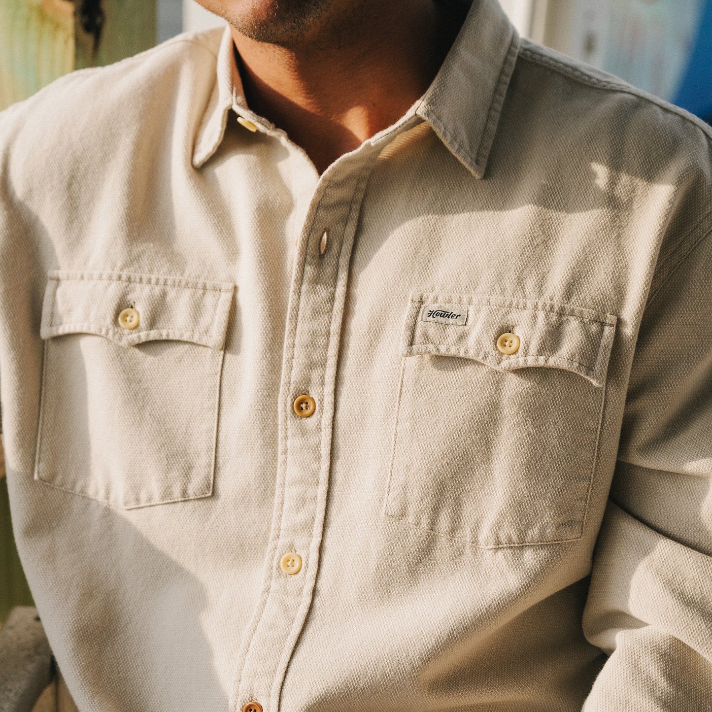 A person wearing the Novato Shirt, a light beige button-up with two chest pockets and a small stitched label on the left pocket, photographed from the shoulders to the waist in natural light.