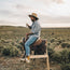 A man in a cotton chambray Tallier Work Shirt, wide-brimmed hat, and jeans sits on a saddle mounted on a wooden frame, holding a drink against open shrubland and a cloudy sky—a timeless nod to classic American workwear.