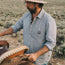 A man in a Tallier Work Shirt made of cotton chambray and a wide-brimmed hat stands outdoors, adjusting a leather saddle on a horse. The scene captures the spirit of American workwear against an open, scrubby landscape.
