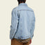 A person with braided hair is shown from the back, wearing a light blue denim jacket with a distinctive lightning bolt stitch over a patterned shirt and brown pants against a light beige background.