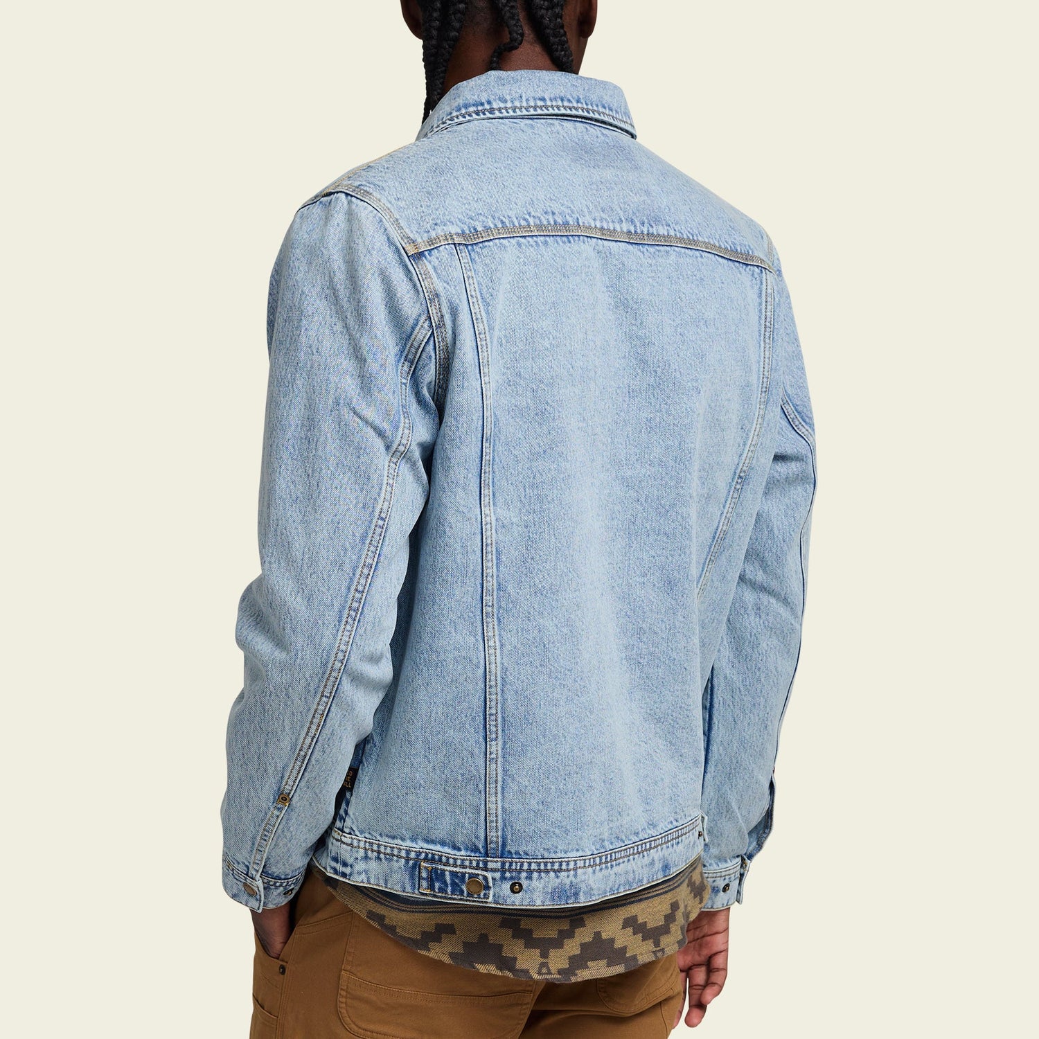 A person with braided hair is shown from the back, wearing a light blue denim jacket with a distinctive lightning bolt stitch over a patterned shirt and brown pants against a light beige background.