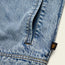 Close-up of light blue denim fabric, likely from a Depot Jacket, showing a vertical pocket with yellow lightning bolt stitch and a small black tag featuring a gold ape face logo on the seam.