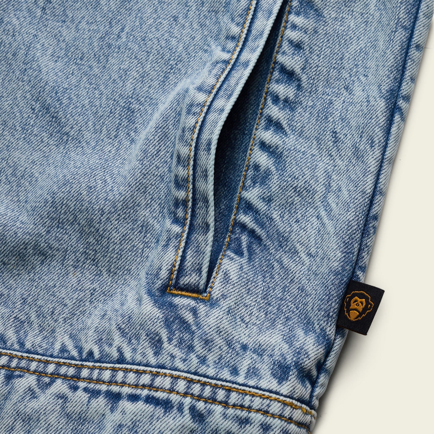 Close-up of light blue denim fabric, likely from a Depot Jacket, showing a vertical pocket with yellow lightning bolt stitch and a small black tag featuring a gold ape face logo on the seam.