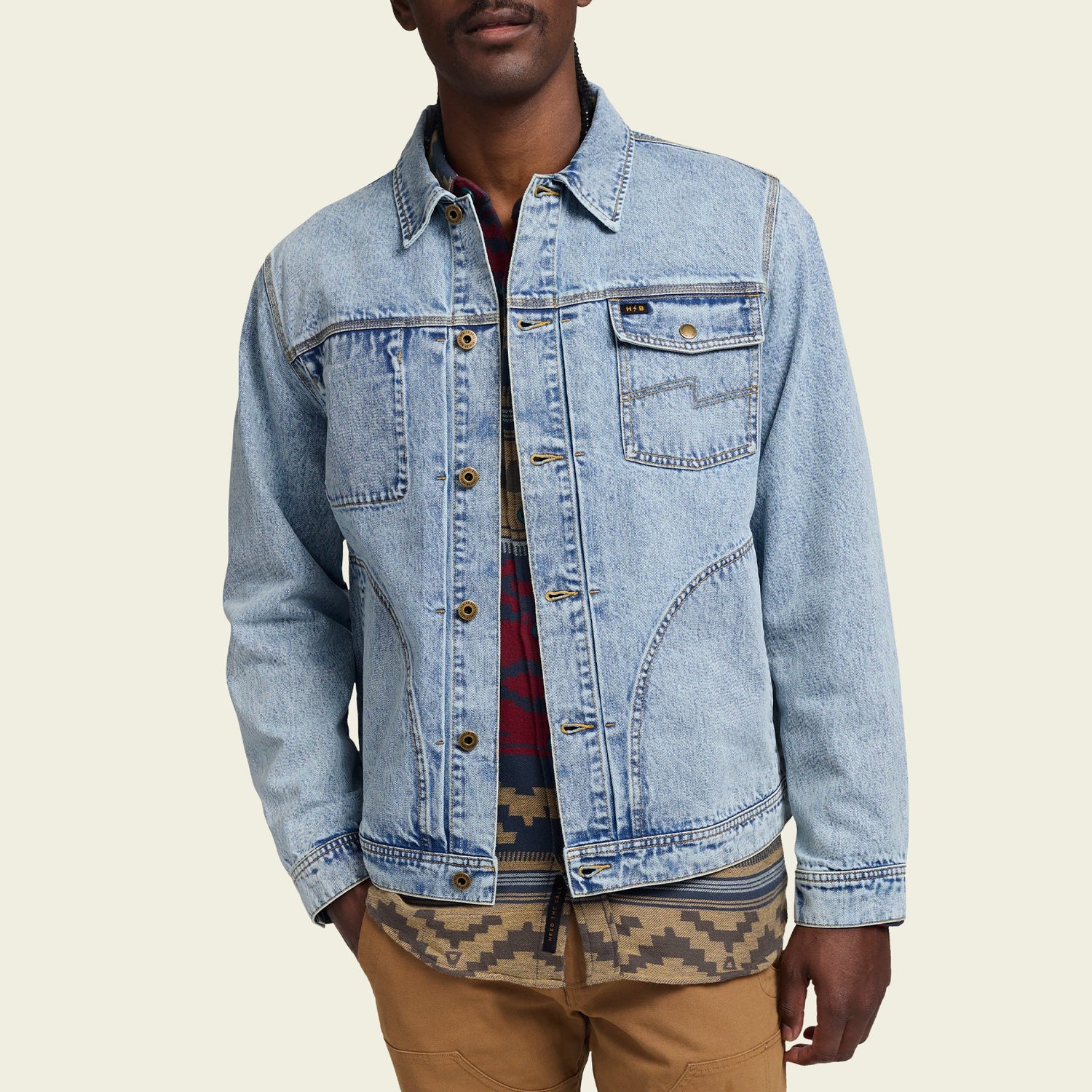 A person wearing a light blue Depot Jacket with lightning bolt stitch over a red and blue plaid shirt and a patterned shirt, paired with tan pants, stands against a plain light background.