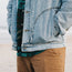 A person stands on the beach with hands in the pockets of a light blue Depot Jacket featuring a subtle lightning bolt stitch, wearing a green patterned shirt underneath and tan pants. The ocean and sand are visible in the blurred background.