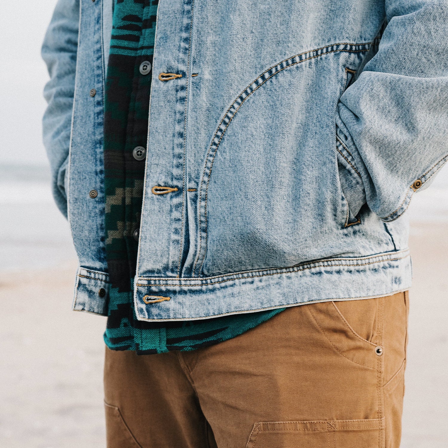 A person stands on the beach with hands in the pockets of a light blue Depot Jacket featuring a subtle lightning bolt stitch, wearing a green patterned shirt underneath and tan pants. The ocean and sand are visible in the blurred background.