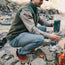 A person in outdoor gear and tech fabric pants kneels on rocks by a river, warming their hands over a portable camping stove with a blue Jetboil cooking system. An orange fuel canister and multitool are nearby.