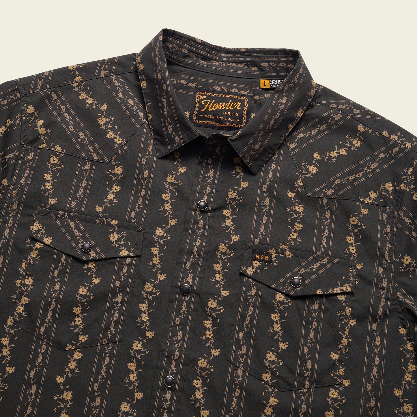 A black western shirt with tan floral and stripe patterns, featuring two front chest pockets with flaps and pearl snaps, plus a woven Howler Bros label on the inside collar.