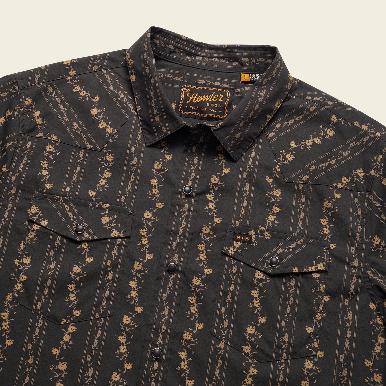 A black western shirt with tan floral and stripe patterns, featuring two front chest pockets with flaps and pearl snaps, plus a woven Howler Bros label on the inside collar.