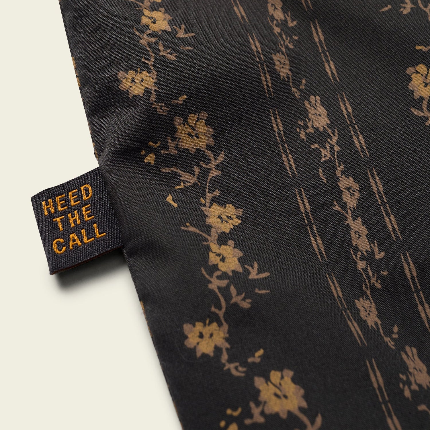 A close-up of a black western shirt fabric with a brown floral pattern and a sewn black tag that reads HEED THE CALL in orange letters, highlighting the distinct style of the H Bar B Snapshirt.