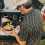 A man in a black western shirt with pearl snaps and a cap holds a skillet of small potatoes beside a truck tailgate, preparing food with raw meat and seasonings laid out on foil.