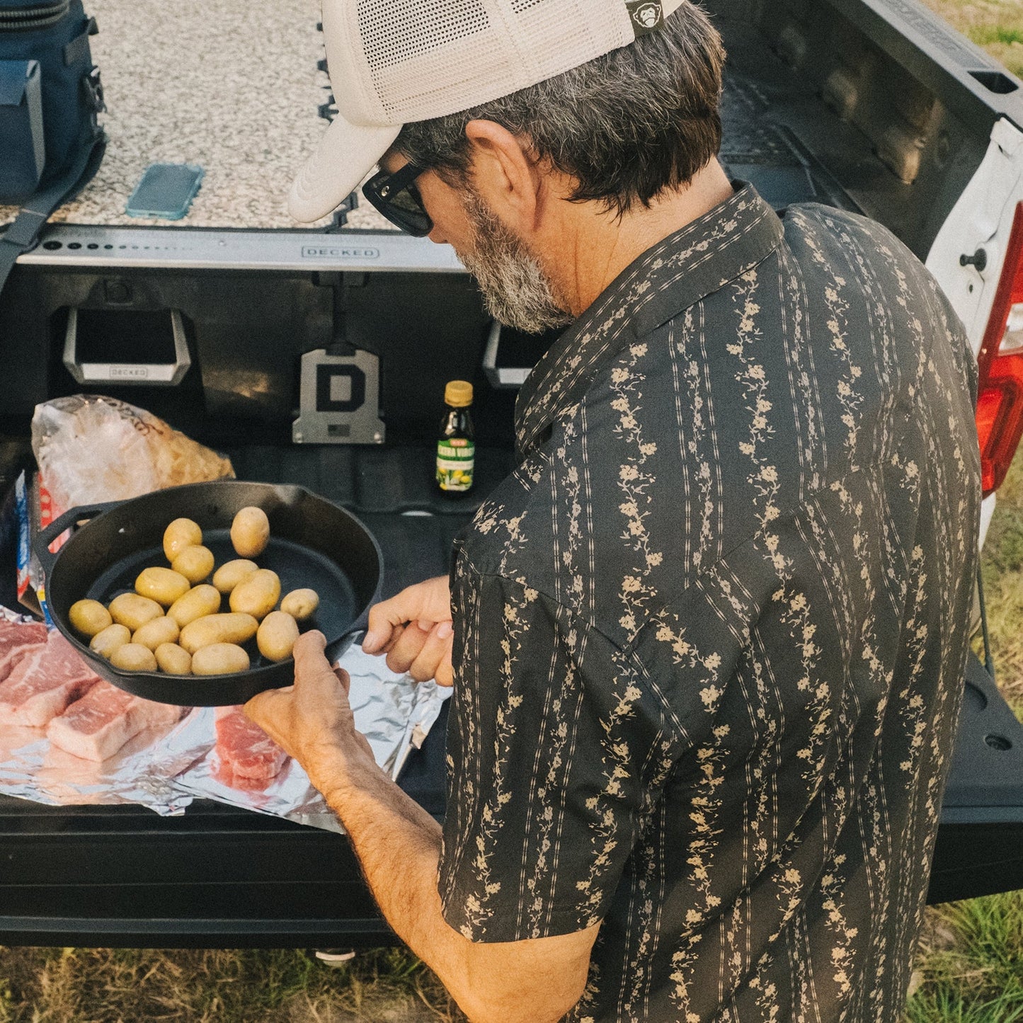 A man in a black western shirt with pearl snaps and a cap holds a skillet of small potatoes beside a truck tailgate, preparing food with raw meat and seasonings laid out on foil.
