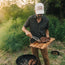 A man in a western shirt and sunglasses uses tongs to place grilled steaks from a wooden board onto a barbecue grill outside, surrounded by tall grass and trees.